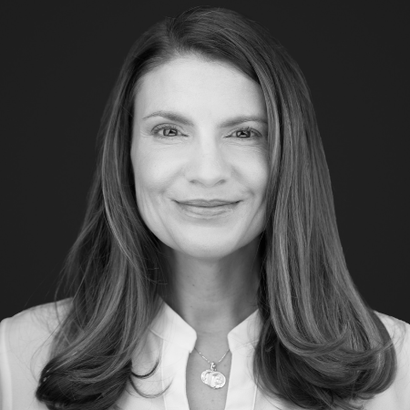 Portrait of a person with long, dark hair wearing a white blouse and a pendant necklace. They have a neutral expression against a dark background. The image is in black and white.