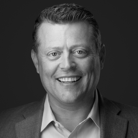 Smiling person with short hair in a suit jacket against a dark background. Black and white portrait.